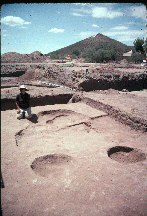 Photograph of a Silverbell interval structure at the Clearwater site. A member of the field crew kneels next to the pit depression to provide scale. She appears to be a woman of average height and the pit is roughly as wide in diameter as she is tall.