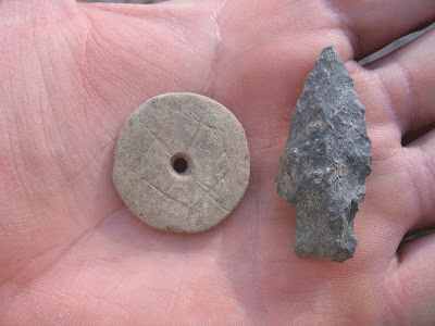 Photo of a San Pedro phase spindle whorl and a San Pedro point from Las Capas resting on a man's palm. The wrhorl is a flat stone disk about the size of a half-dollar coin with a hole in the center. Four incised lines extend from the hole in a + design, and four more incised lines form a square centered on the hole. he point is about as long as the man's pinky finger.