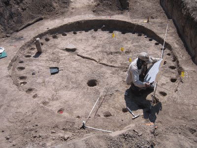 Photo of Allen Denoyer mapping a Cienega phase big house. Allen crouches near the side of a large round pit that is roughly 10 ft in diameter. multiple closely spaced postholes are arranged around the inner perimeter of the shallow pit. A foot-high rectangular stone bar stands at the side of the pit opposite Allen.
