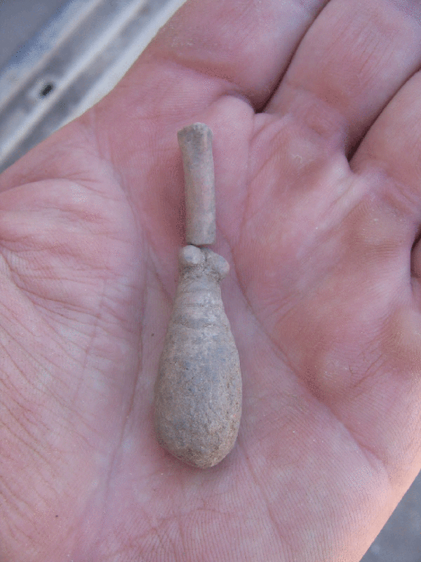 Photo of a fired-clay figurine from Las Capas, resting on a man's palm. The shape is an extended teardrop, with a long, narrow bulb-shaped base and a very narrow cylindrical top. At the juncture of the two segments are two small balls that have been interpreted as female breasts.. The figurine is roughly as long as the man's pinky finger.
