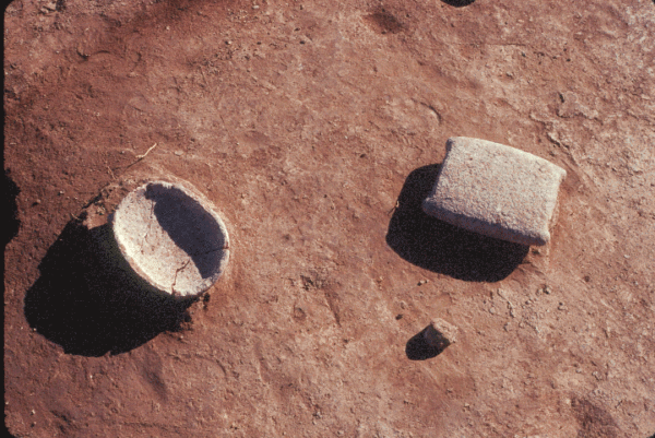 Photo of two ground stone trays on the floor of a Cienega phase pithouse. The one on the left is oval-shaped and right side up. The one on the right is subrectangular, with spurs or barbs at the corners, and is upside down. Each is roughly the size of a large adult male hand.