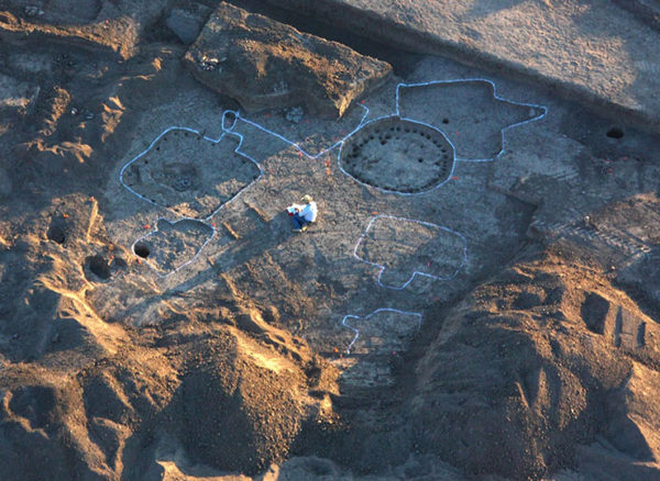 Aerial photo of Homer Thiel drawing a map of an excavated pit structure. He is wearing a white shirt and straw hat, working on a n oversized clipboard