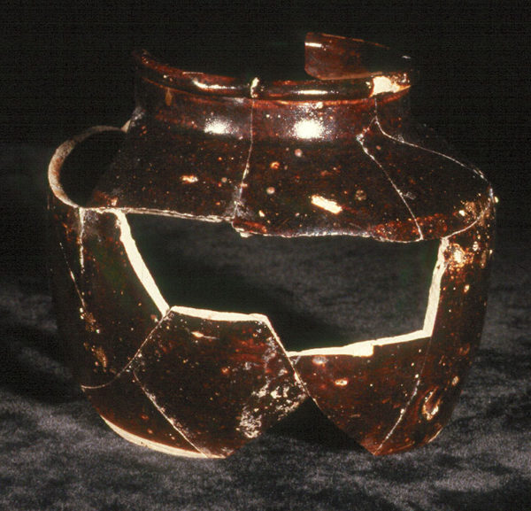 Photo of a shattered wide-mouth ceramic jar that has been glued together, although several pieces are missing. It is short and squat with a wide mouth, is dark brown, and may have held fermented or pickled foods.