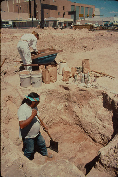 Old photo of the Desert field crew excavating a privy. A man with a shovel stands in the pit, while another man off to the side examines a screen set over a wheelbarrow.