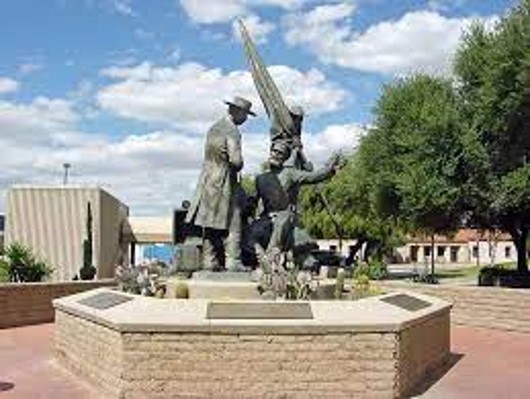 Photo with a rather distant view of a statue in downtown Tucson showing two members of the Mormon Battalion trading with Teodoro Ramirez. One of the Mormon figures has a flat-brimmed hat. The other is holding a furled flag. The statue is on a six-sided adobe base with a concrete cap. Fluffy clouds are in the sky.