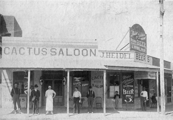 Photo taken in 1900 of the Cactus Saloon exterior. The saloon keeper is standing outside with a white apron, flanked by four men to his sides. Large painted signs advertise 5-cent beer.
