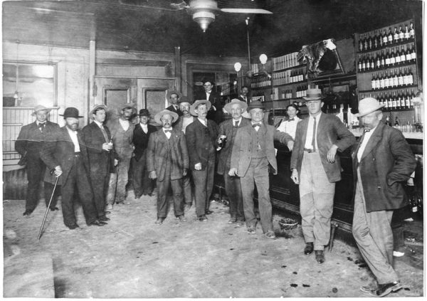 Historical photo of the Cactus Saloon interior. Twelve men stand around the bar, wearing jackets, waistcoats, and hats. The bartender has a white shirt and black bowtie. A leopard pelt is draped over the mirror behind the bar.