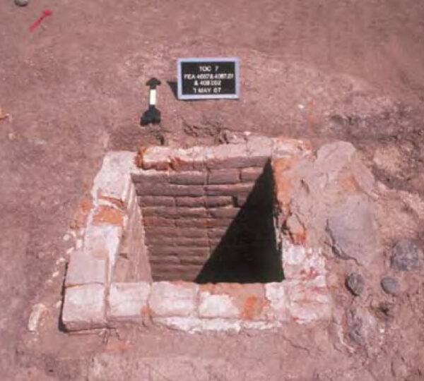 Photo of Feature 4087, the brick-lined well. It is a nearly square hole in the ground with walls made of dusty red flat adobe bricks. 