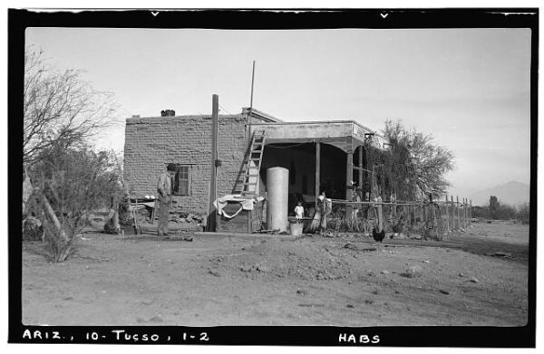 Black-and-white photo of the Carillo house, taken in 1938, showing an adobe structure with a covered wooden porch. A man stands in the left part of the frame. A small child stands on the porch.
